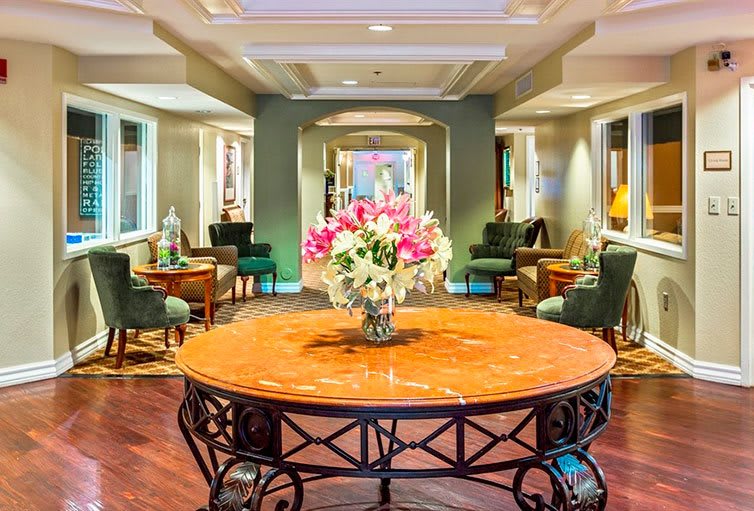 A brightly lit hotel lobby featuring a central table adorned with a bouquet of flowers, surrounded by comfortable seating.