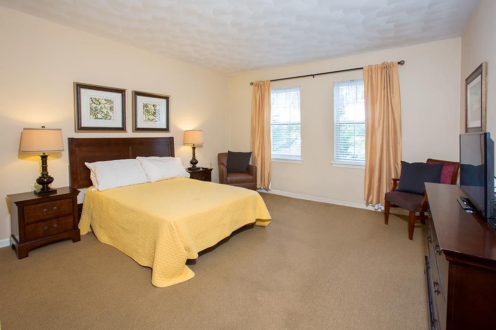 A well-decorated bedroom featuring a bed with a yellow cover, two chairs, decorative art on the wall, and windows with curtains.