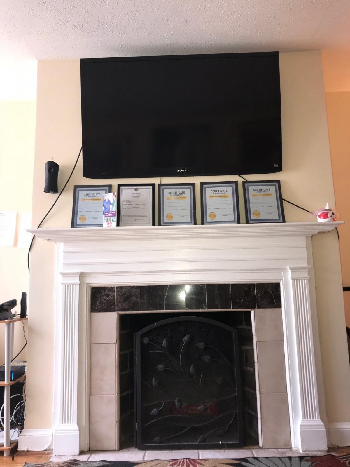 A living room with a large television mounted above a white fireplace mantel, which is decorated with several framed plaques and small items.