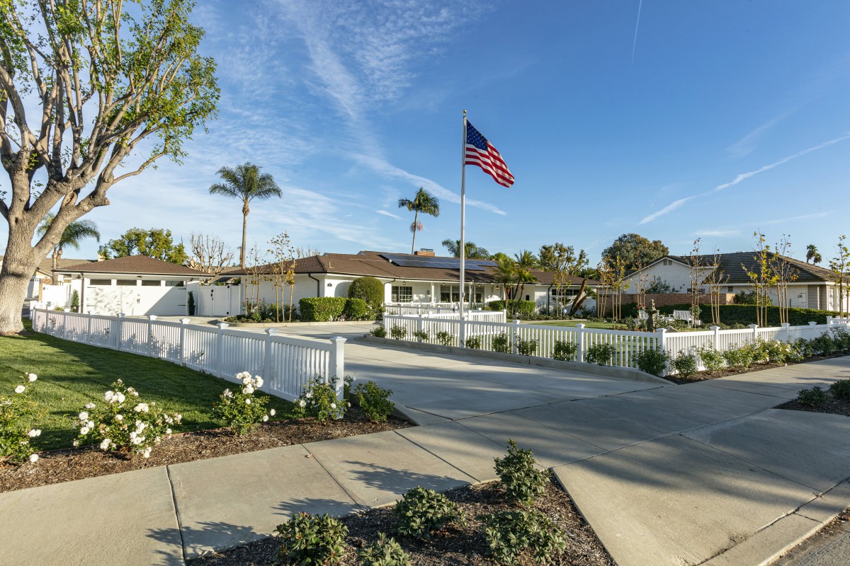 A suburban house with a driveway, featuring an American flag and palm trees under a clear blue sky.