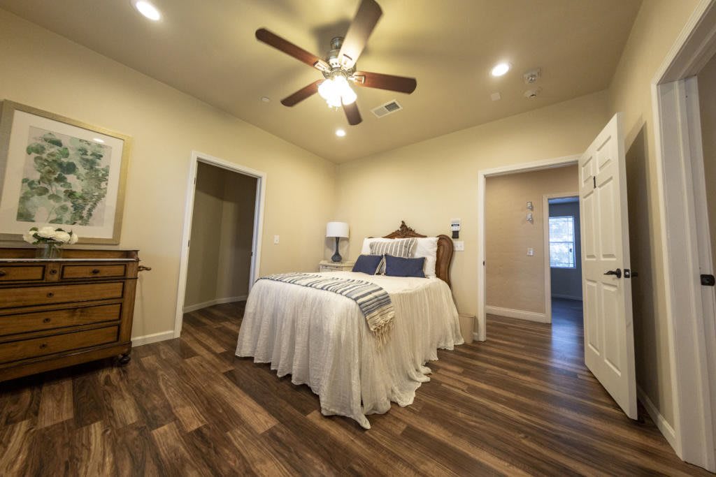 A cozy bedroom with a wooden floor, featuring a comfortable bed and ceiling fan.