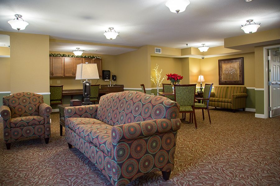 A cozy living room with patterned sofas, a dining table and chairs, warm lighting, and carpeted floors.
