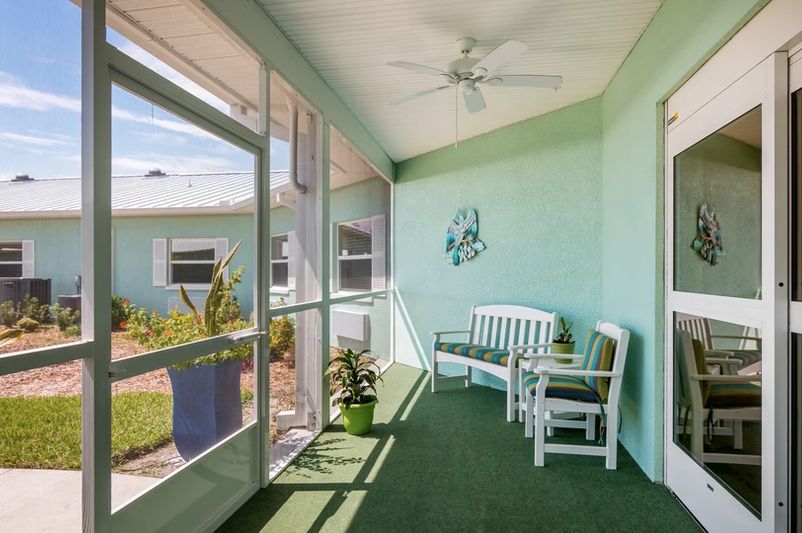 A bright, screened porch with green walls, featuring bench seating and indoor plants, complemented by a ceiling fan above.