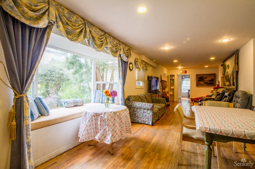 A cozy living room with wooden floors, decorated with floral curtains and a sofa. A dining table is set near a large window offering a view of the garden.