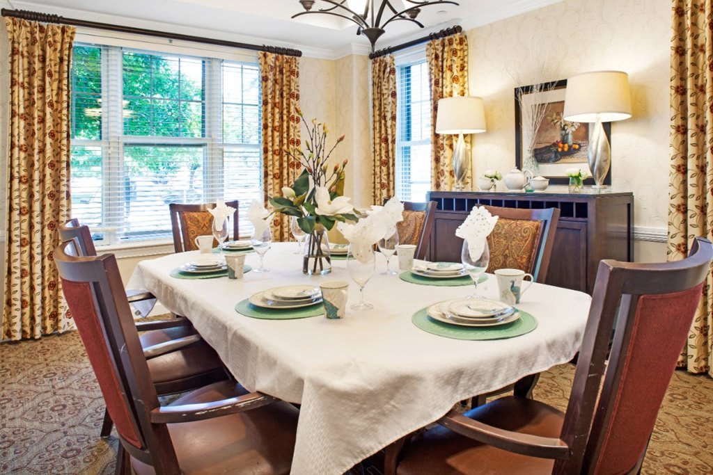 An elegantly set dining room table with a white tablecloth and green accents, surrounded by wooden chairs and featuring large windows with patterned curtains.