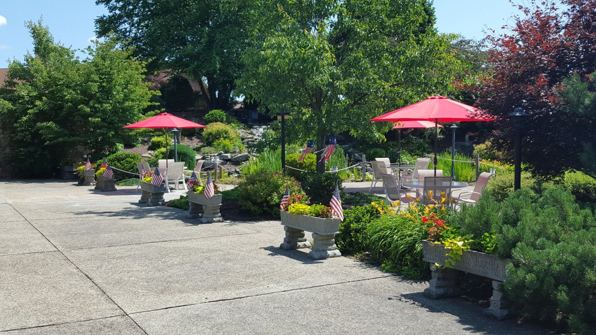 A sunny outdoor garden patio with red umbrellas, surrounded by lush greenery and colorful flowers.