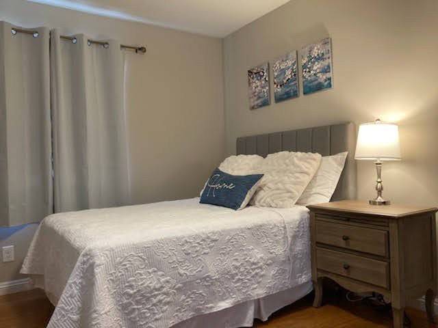 A cozy bedroom with a neatly made bed, featuring white bedding and decorative pillows, accompanied by a nightstand with a lamp and wall art above the headboard.
