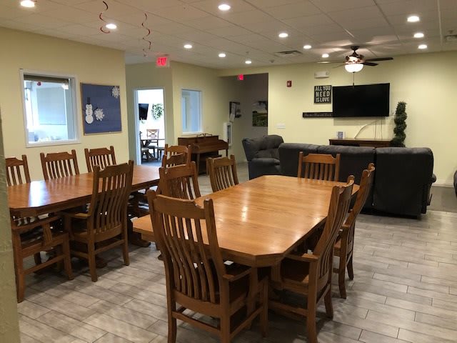 A spacious meeting room with a large wooden table, multiple chairs, a wall-mounted TV, and a ceiling fan.