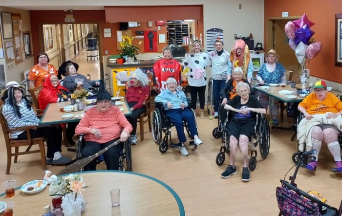 A group of elderly people in wheelchairs poses for a photo indoors, some wearing costumes.