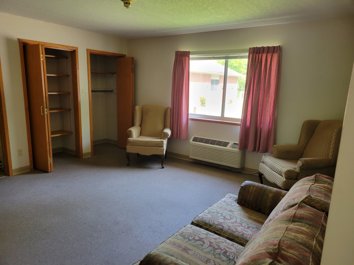 A living room with a sofa, two chairs, a window with curtains, and empty wooden shelves.