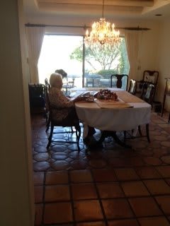 A dining room with two people sitting at a table, illuminated by natural light from a large window.
