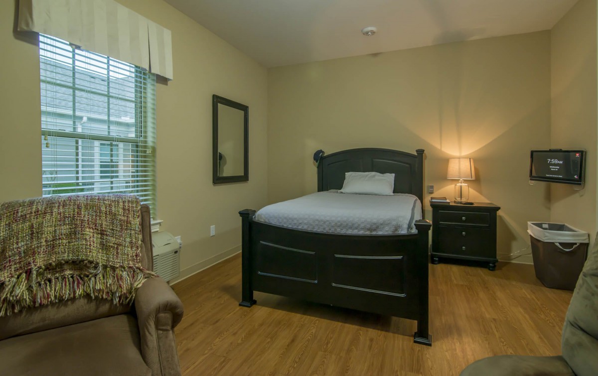 A cozy bedroom with a single bed, nightstand, and chair, featuring hardwood floors and soft lighting.