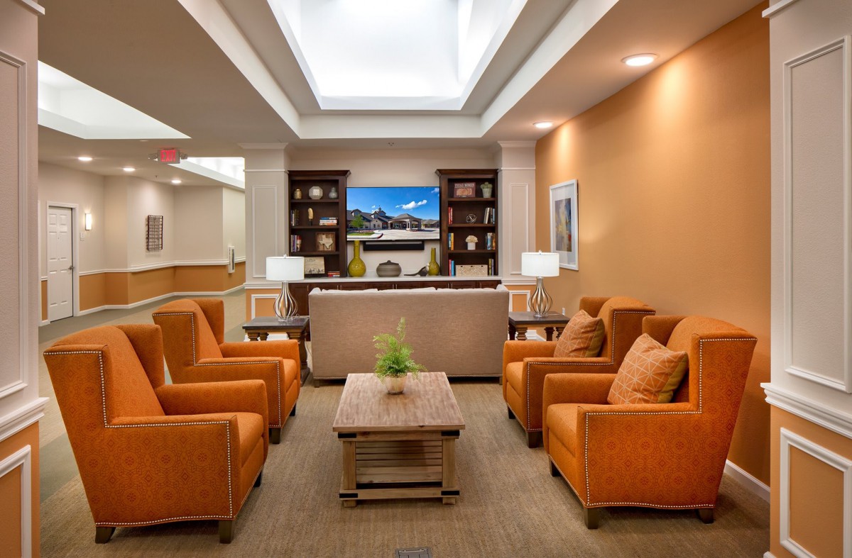 A modern lounge area featuring orange chairs, a central coffee table, and a bookshelf against one wall, all under bright lighting.