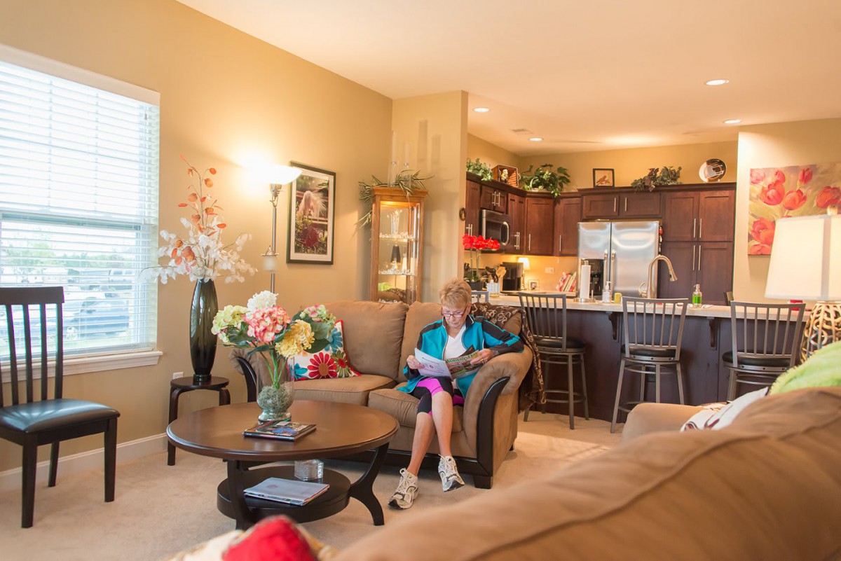 A cozy living room with a woman reading on a sofa, surrounded by decor and flowers, with an open view of the kitchen.