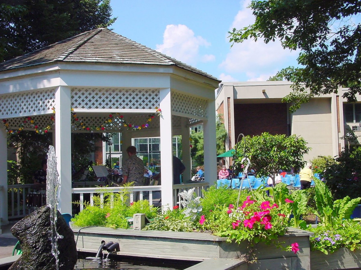 A picturesque gazebo surrounded by vibrant flowers and lush greenery near a small pond.