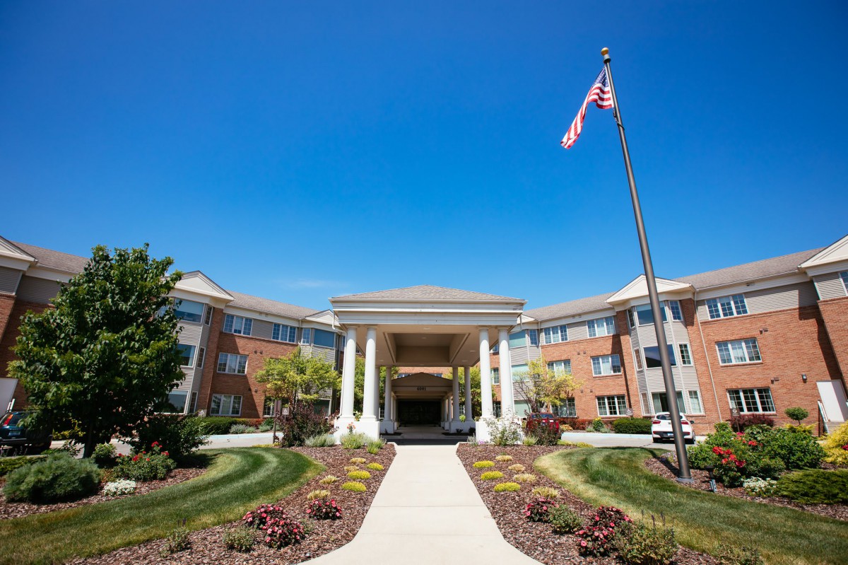 A large building with a grand entrance, surrounded by landscaping, under a clear blue sky with a flagpole in front.