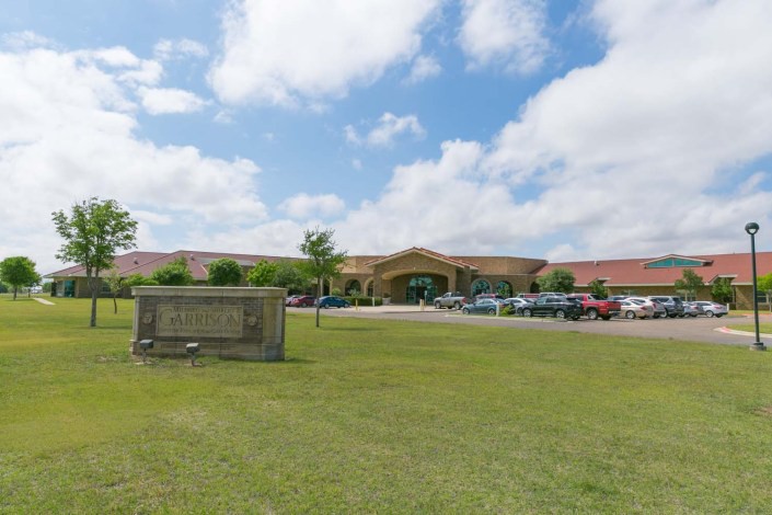 A school building with a parking lot, surrounded by grass under a partly cloudy sky.