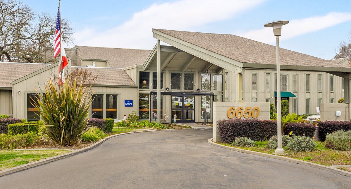 The image shows the entrance of a contemporary building with a driveway and landscaped surroundings.