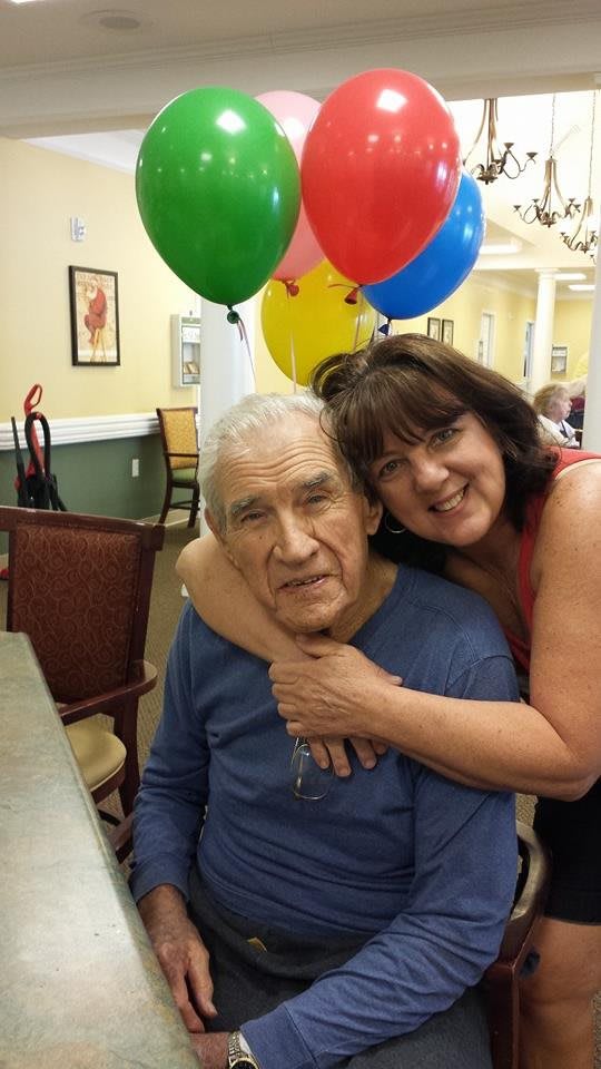 A woman embraces an elderly man with colorful balloons in the background, suggesting a celebration or special occasion.