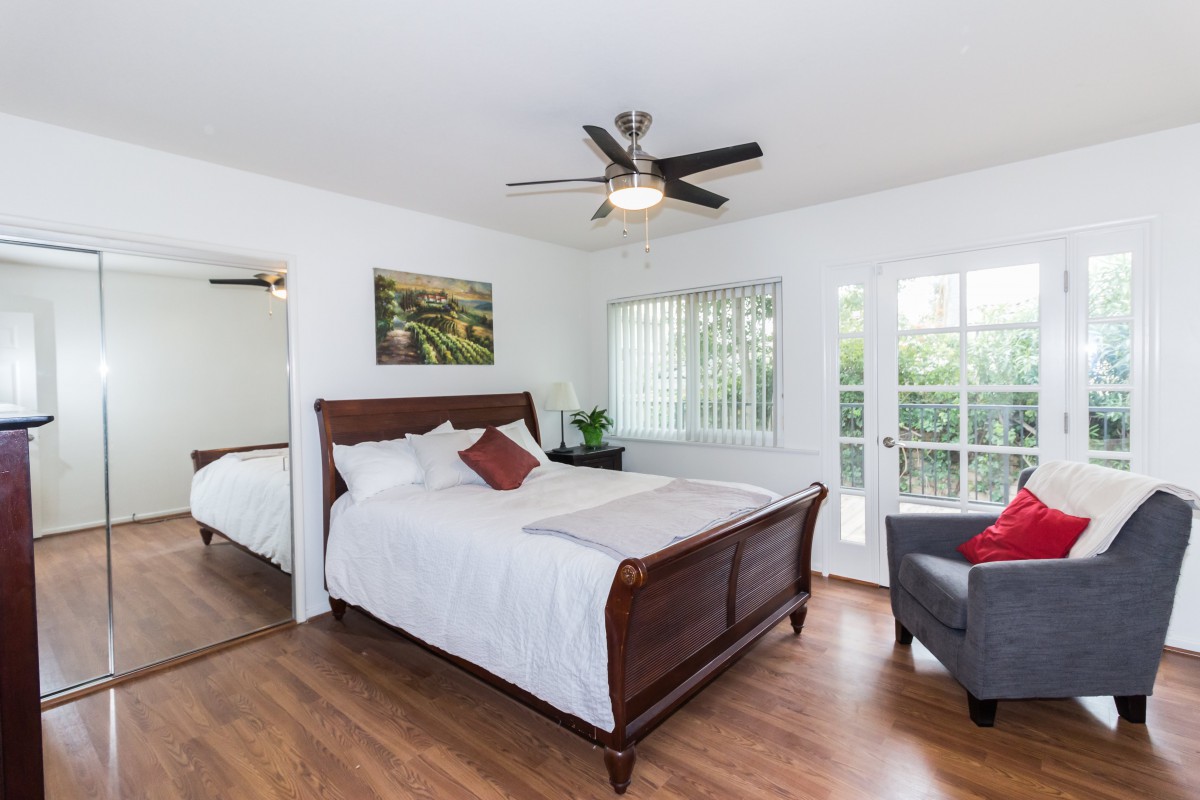 A cozy bedroom with a wooden bed and a ceiling fan, featuring bright natural light from the windows and glass doors.