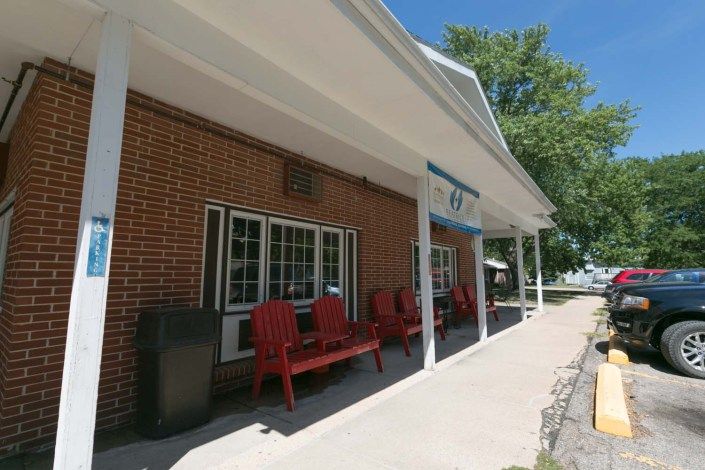 A brick building with a porch featuring red chairs and an awning on a sunny day.