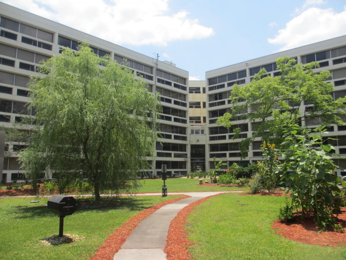 An outdoor area with a central pathway leading to a modern multi-story building, surrounded by lush greenery and trees under a partly cloudy sky.