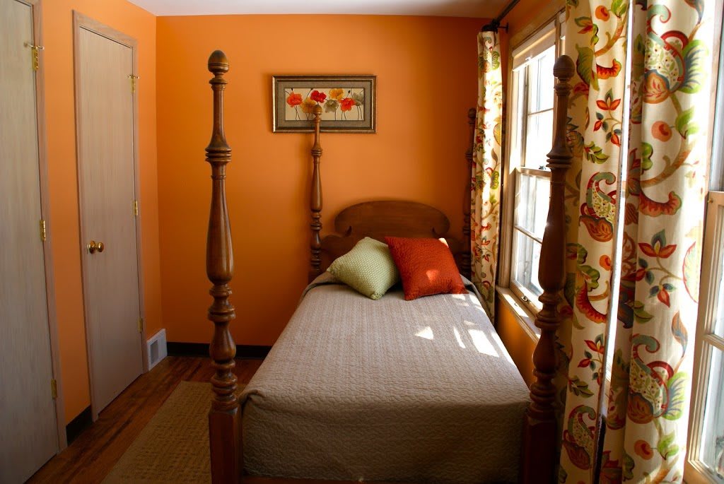 A cozy bedroom with orange walls features a twin bed adorned with colorful decorative pillows and floral curtains framing the window.