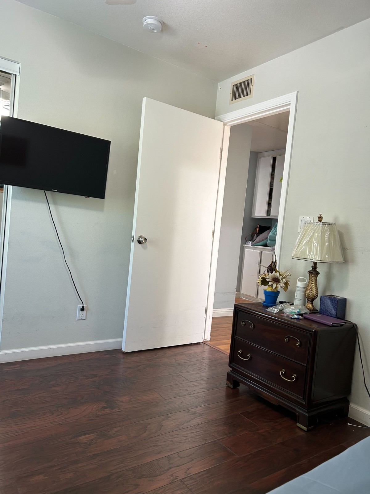 A cozy bedroom with an open door, wall-mounted television, and a dresser with a lamp.