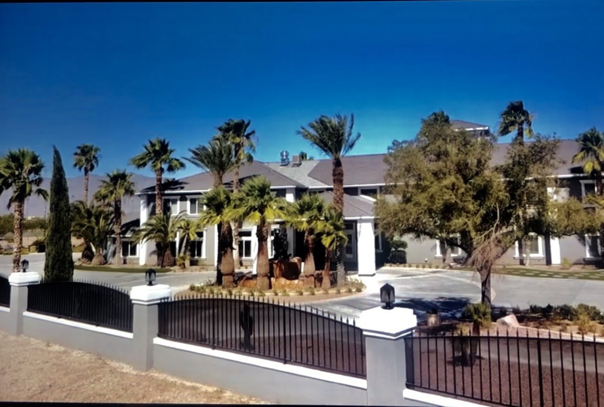 A luxurious mansion surrounded by palm trees and enclosed by a gated fence.