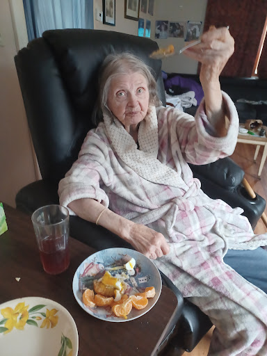 An elderly woman in a robe enjoying a snack while sitting comfortably.
