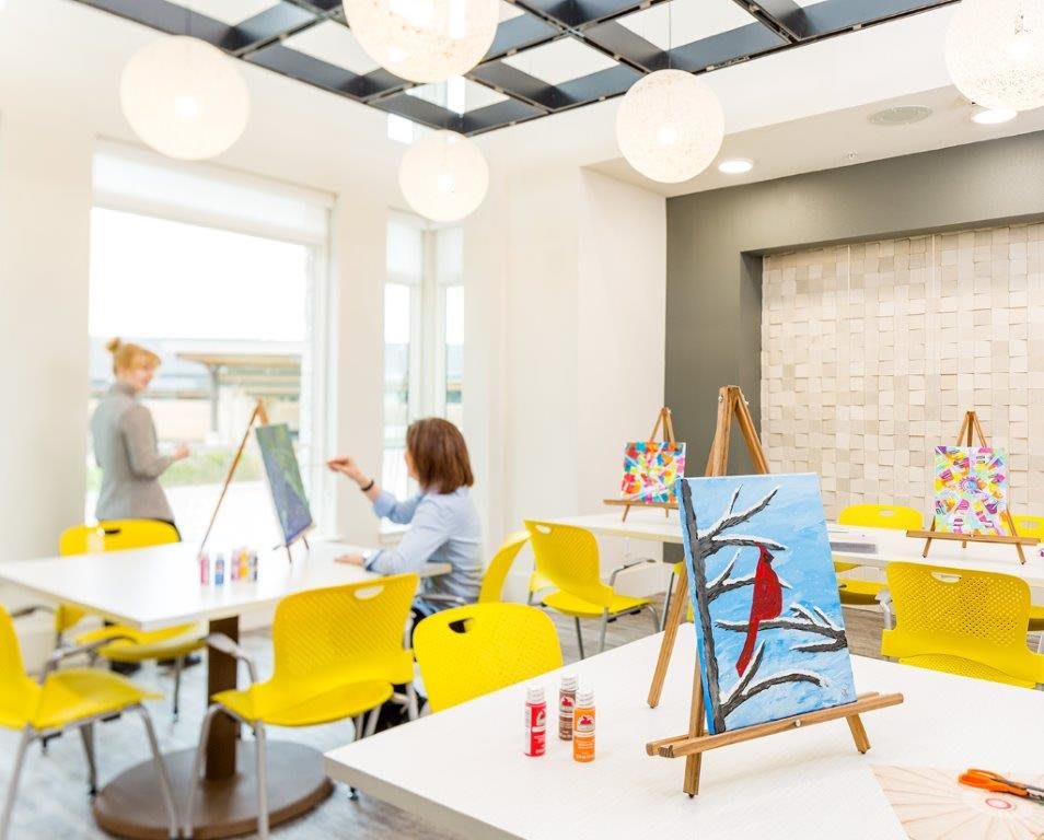 An art class in session with people painting on easels in a brightly lit studio with yellow chairs.