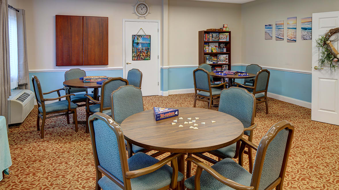 A community room featuring several tables with chairs, a puzzle in progress, and a bookshelf filled with games.