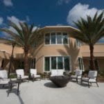 Luxurious house with a spacious patio, featuring palm trees and outdoor seating around a fire pit under a clear blue sky.