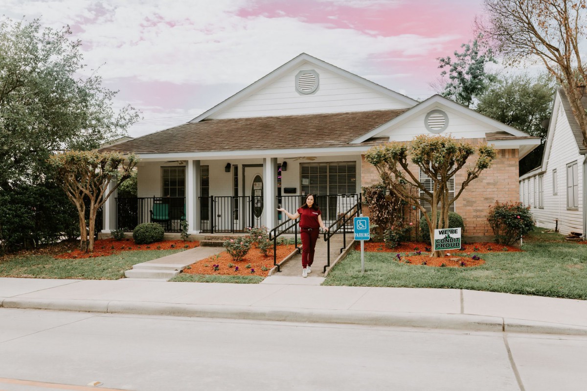 A person stands in front of a house with a porch, surrounded by landscaping and trees.
