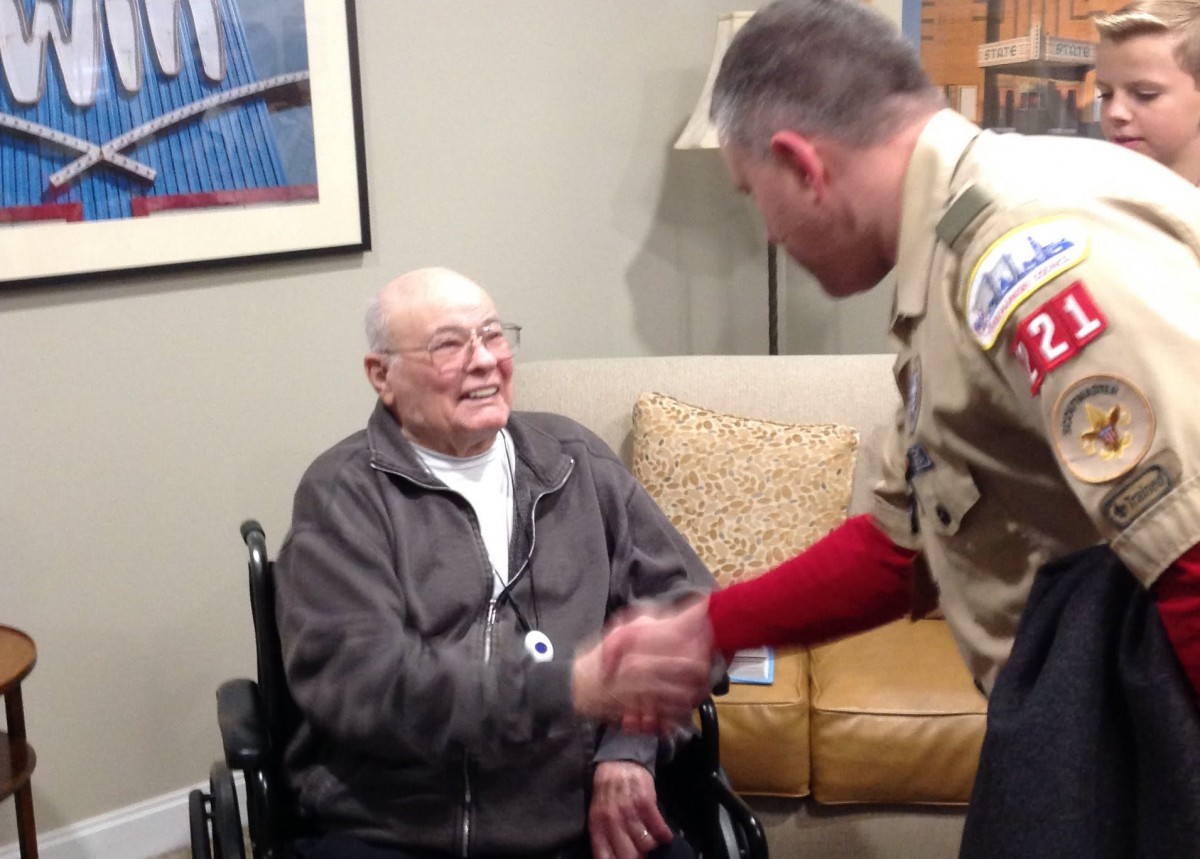 An elderly person in a wheelchair greets someone in a uniform with a handshake indoors.