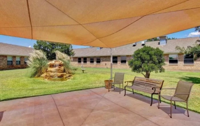 A cozy patio area with a shade sail, a bench, and a view of a garden fountain.