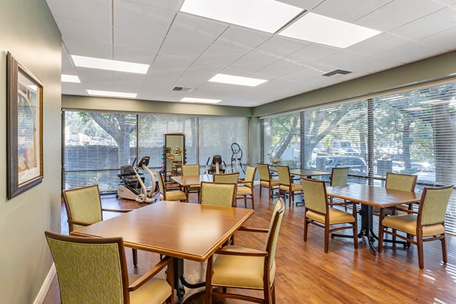 A bright and spacious room featuring dining tables, chairs, exercise equipment, and large windows.