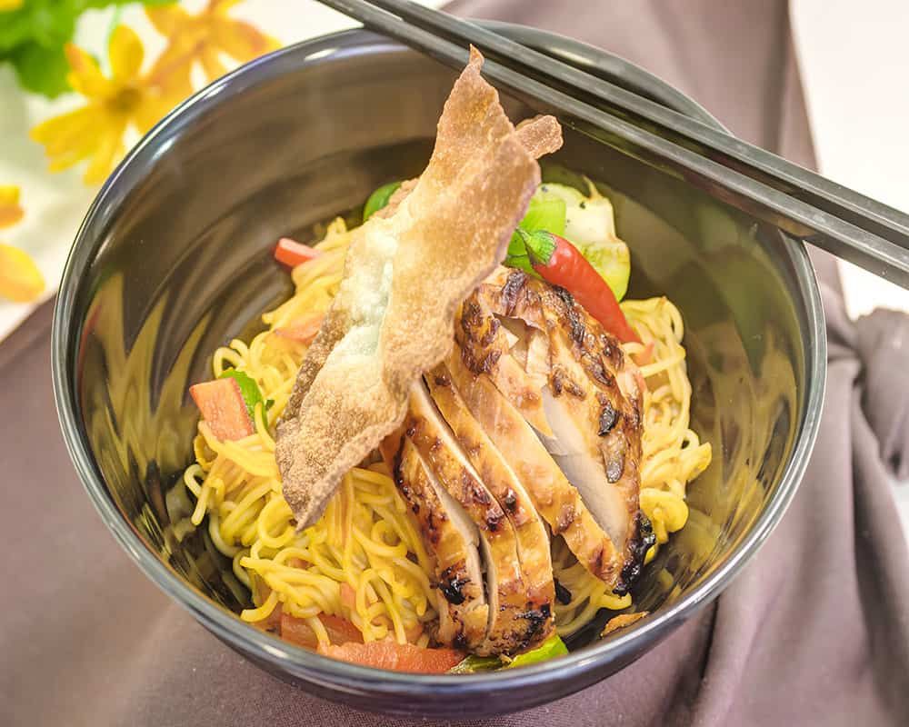 A bowl of noodles topped with grilled chicken and vegetables, garnished with a crispy piece and paired with chopsticks.