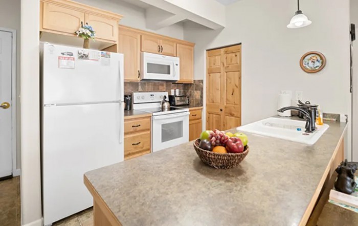 A modern kitchen with wooden cabinets, featuring a refrigerator, stove, microwave, and a fruit bowl on the countertop.