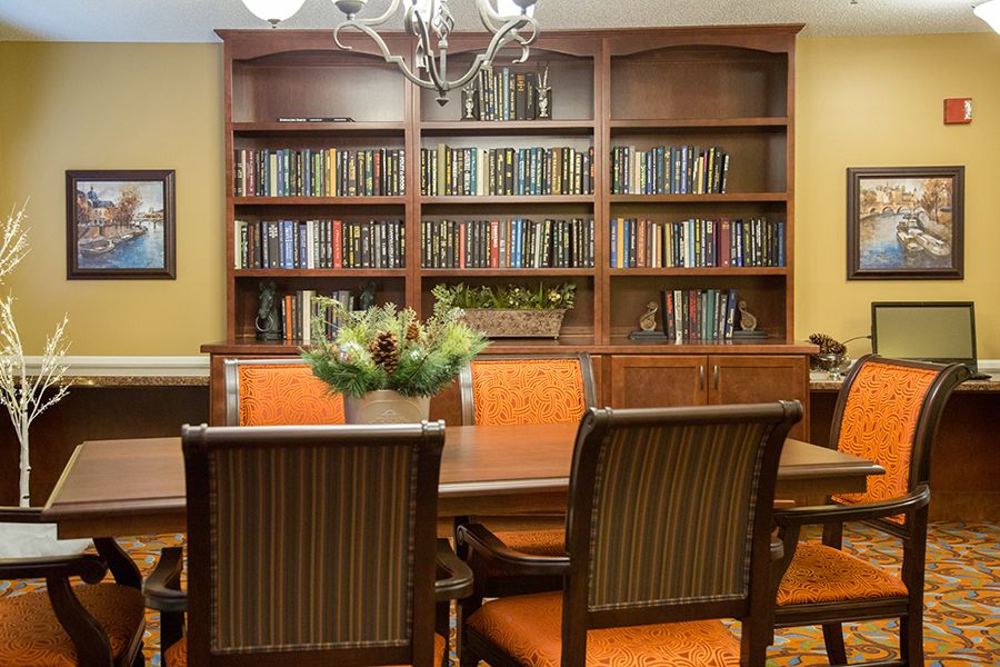 A cozy library setting with wooden bookshelves, a central table surrounded by chairs, and a chandelier overhead.