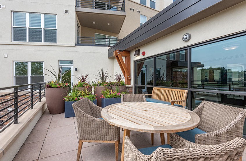 A modern outdoor patio with a round wooden table and wicker chairs surrounded by potted plants in an urban apartment setting.