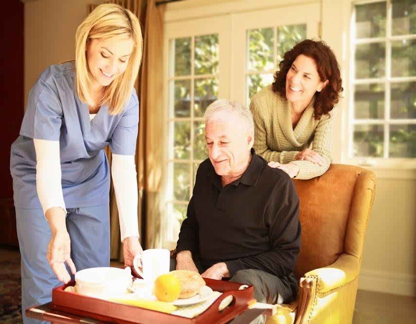 A caregiver assists an elderly man at home while a woman looks on.