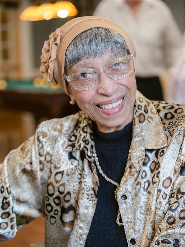 An elderly woman smiles warmly, wearing glasses, a headscarf, and a patterned shirt.