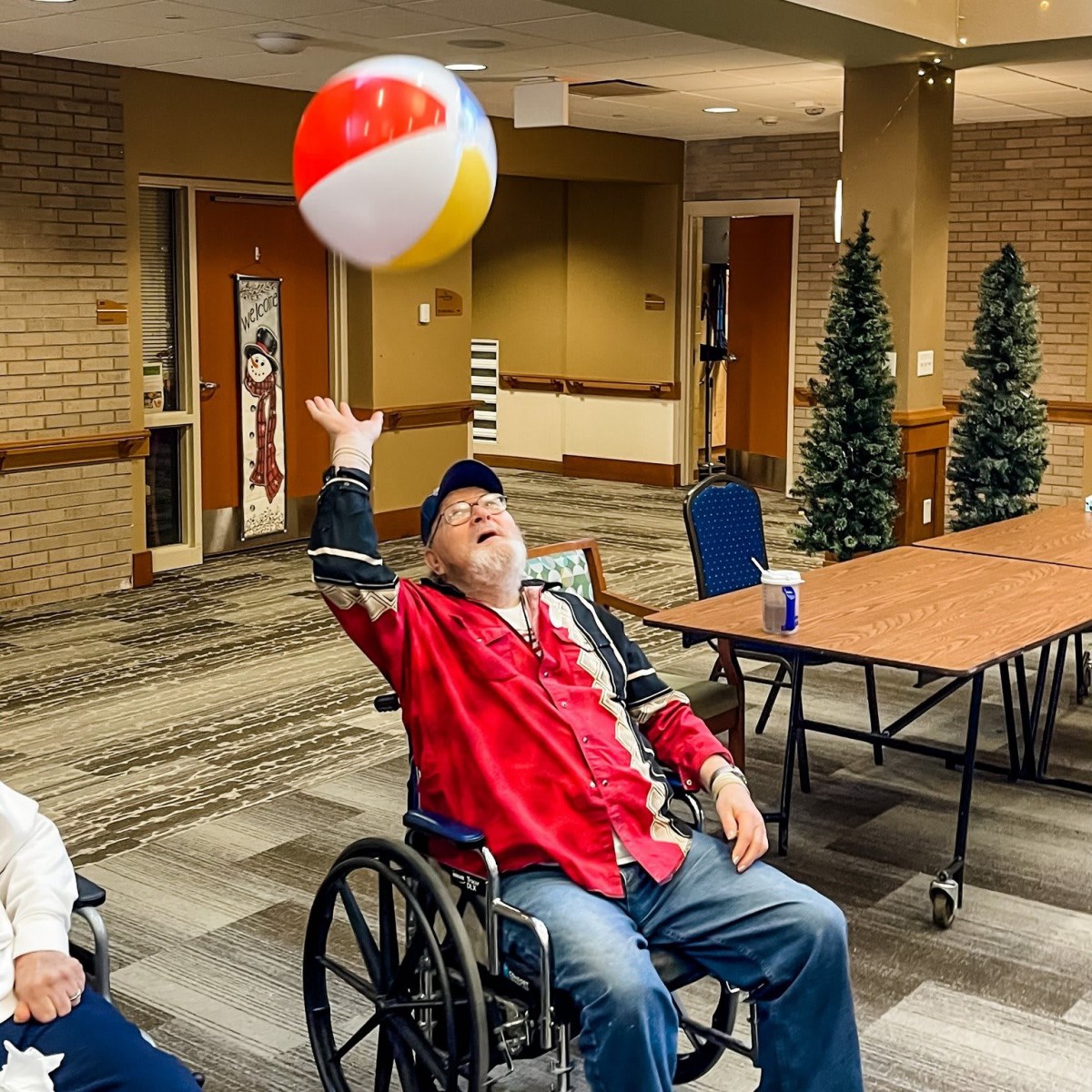 An elderly man in a wheelchair joyfully plays with a beach ball indoors.