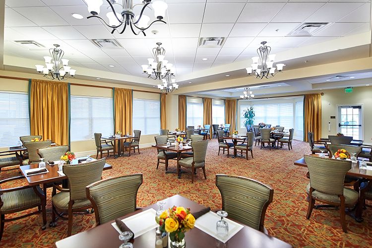 A spacious and elegantly decorated dining room with multiple tables and chandeliers, featuring large windows and a warm carpeted floor.