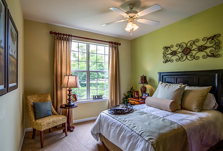 A cozy bedroom with a bed, chair, and lamp, illuminated by natural light from a large window.