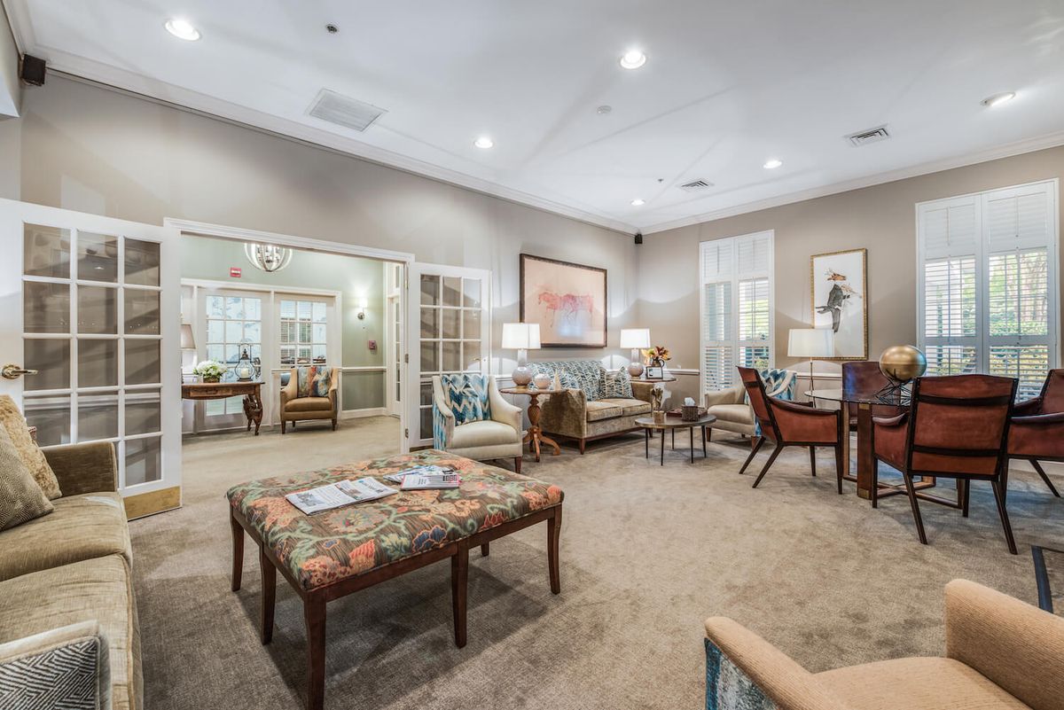 A spacious and elegantly decorated living room featuring a sofa, chairs, tables, lamps, and artwork on the walls.