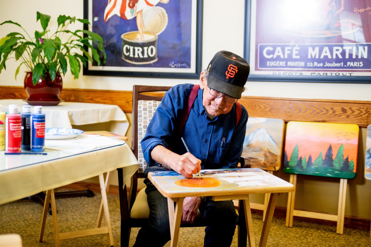 An artist wearing a cap is focused on painting while seated indoors.