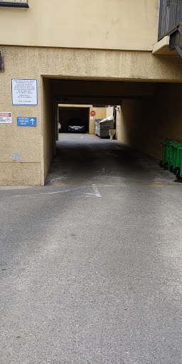 An urban alleyway leading to a parking area, with green garbage bins lined up on the right.