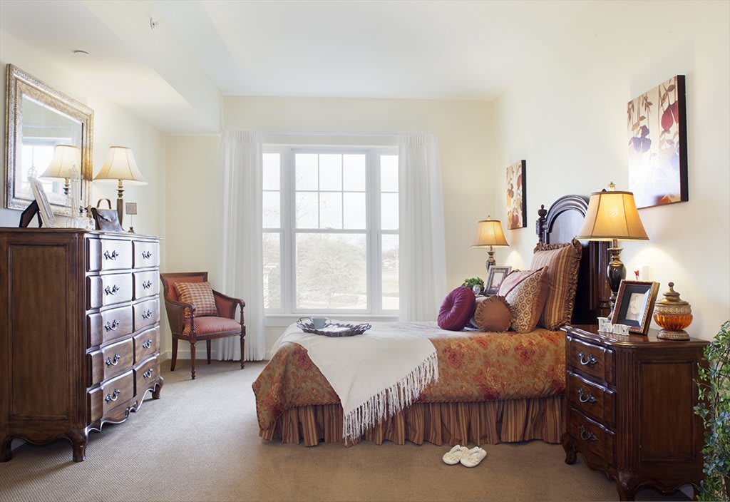 A cozy bedroom with classic furniture, featuring a bed, armchair, dressers, and a large window.